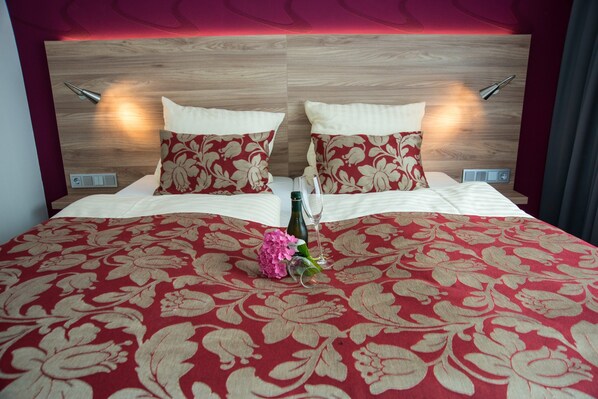 A hotel room with a wooden headboard, two bedside tables, a red floral bedspread, and a vase with flowers. - Hotel Hellers Krug (Holzminden)