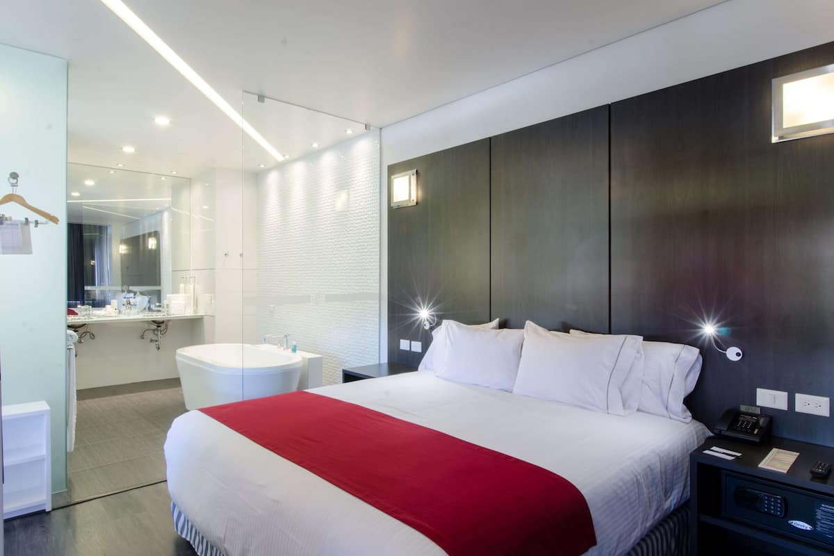 A modern hotel room with a large bed, a bedside table with a lamp, a built-in shelf, and a separate bathroom with a mirror and sink.