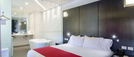 A modern hotel room with a large bed, a bedside table with a lamp, a built-in shelf, and a separate bathroom with a mirror and sink.