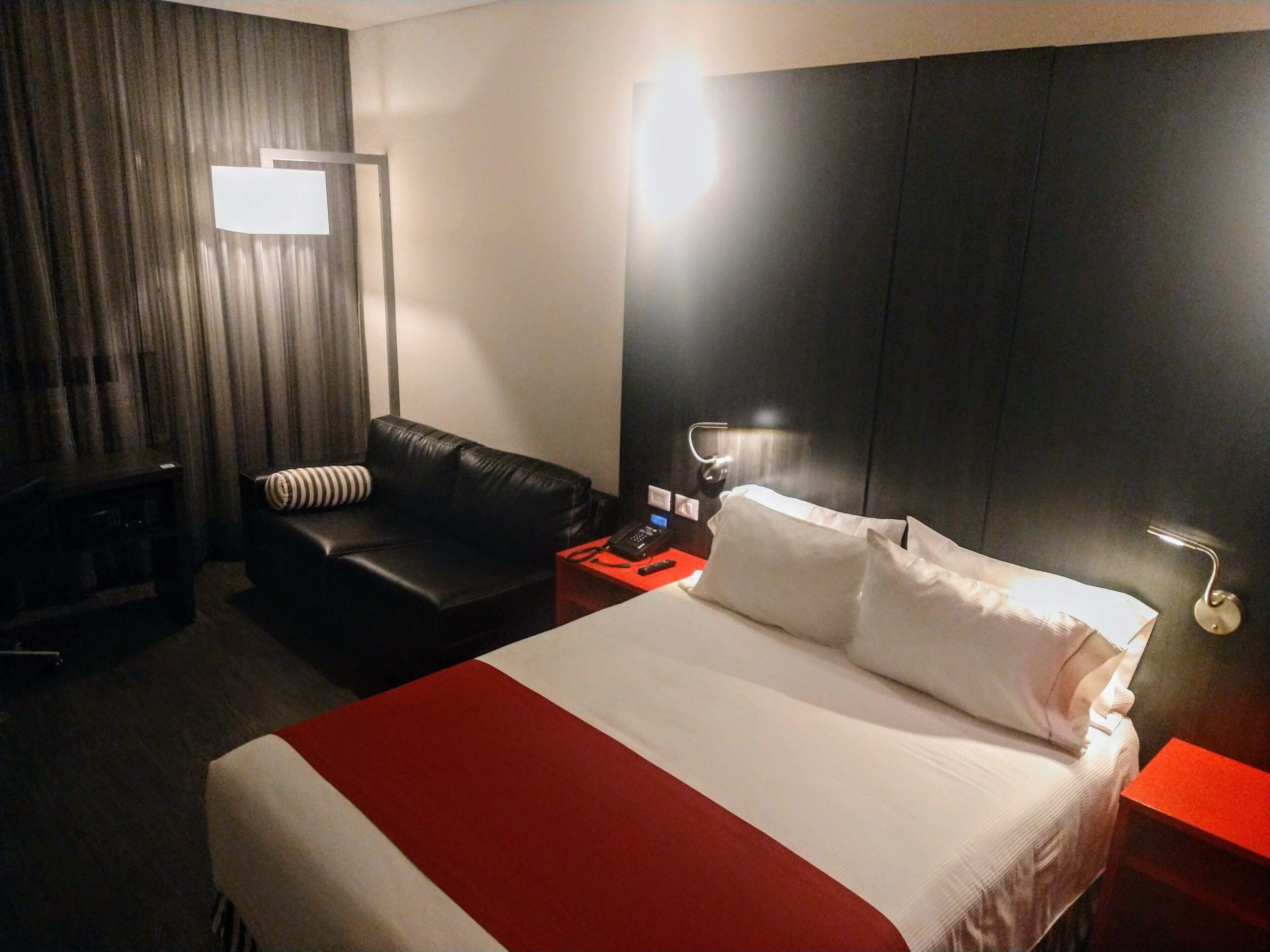 Een hotelkamer met een groot bed, een zwarte leren bank, een rode bijzettafel en een aan de muur bevestigde televisie.