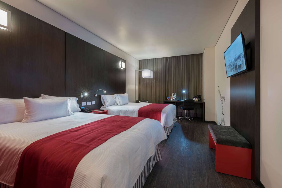A hotel room with two beds, a desk, a TV, and a bench.