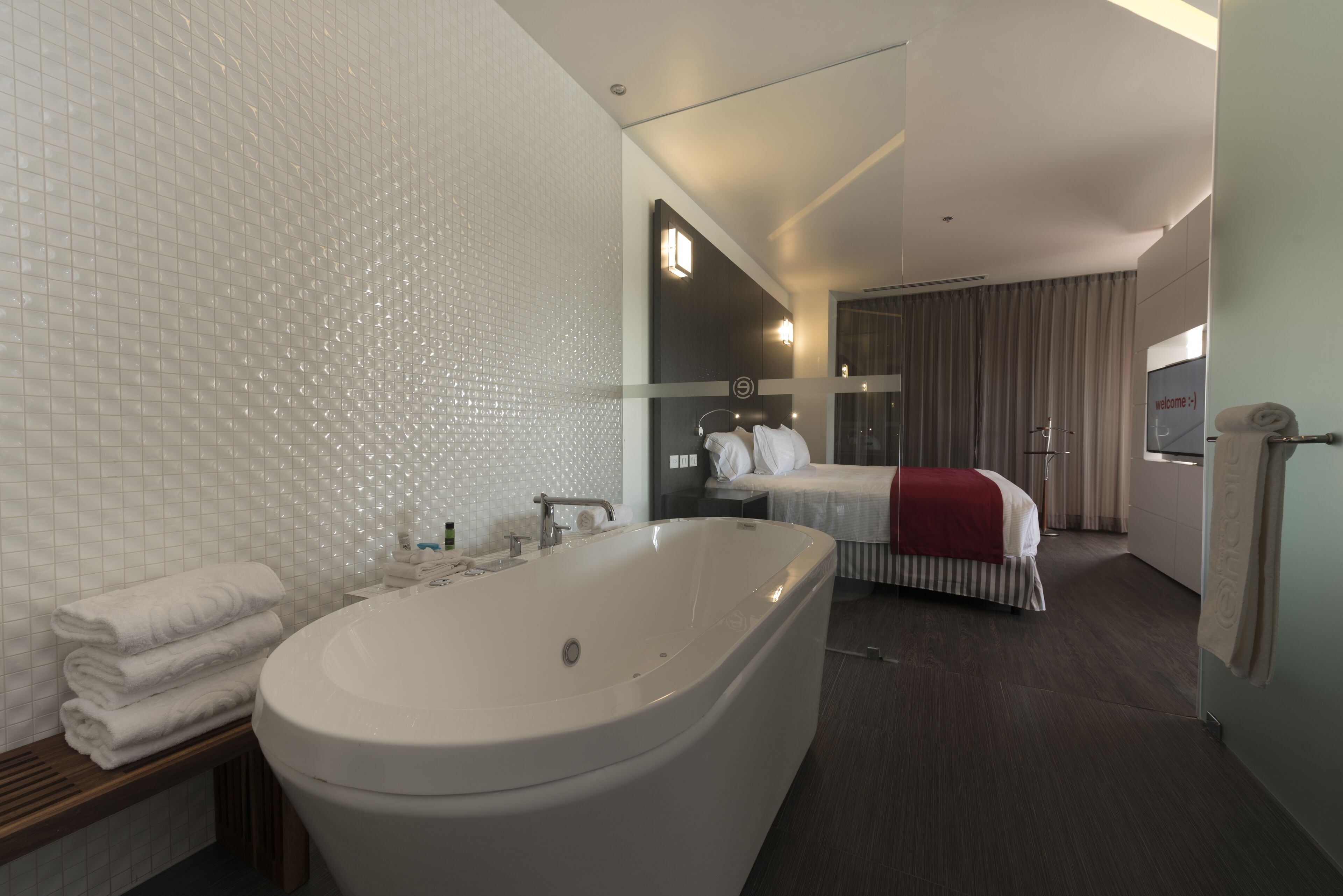 Ein modernes Badezimmer mit einer großen Badewanne, einer Toilette und einer Duschkabine. Ein Bett mit roter Decke und ein Nachttisch mit Lampe.