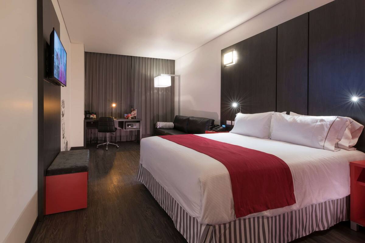 A modern hotel room with a large bed, a desk, a chair, a TV, and a bedside table.