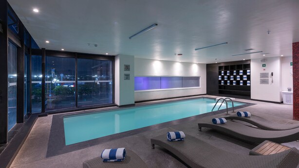 A modern indoor pool area with lounge chairs and a view of the city at night. - Ramada Encore by Wyndham Aguascalientes (Aguascalientes)