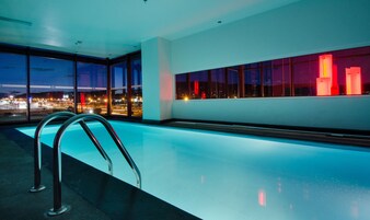 Una piscina interior moderna con paredes de vidrio transparente, una escalera metálica y vista a un paisaje urbano nocturno.
