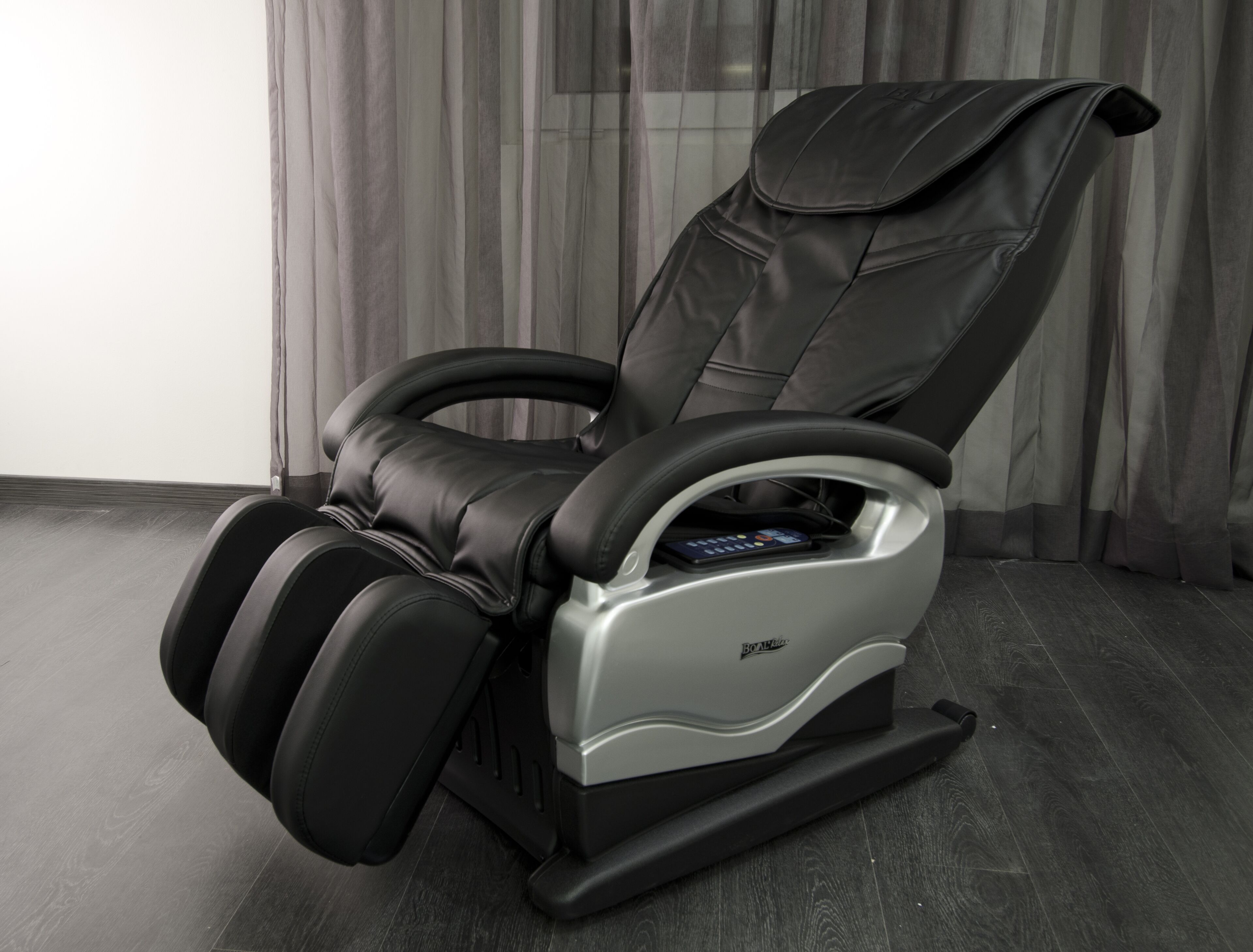 A massage chair with a control panel, positioned in a room with a dark wooden floor and sheer curtains.