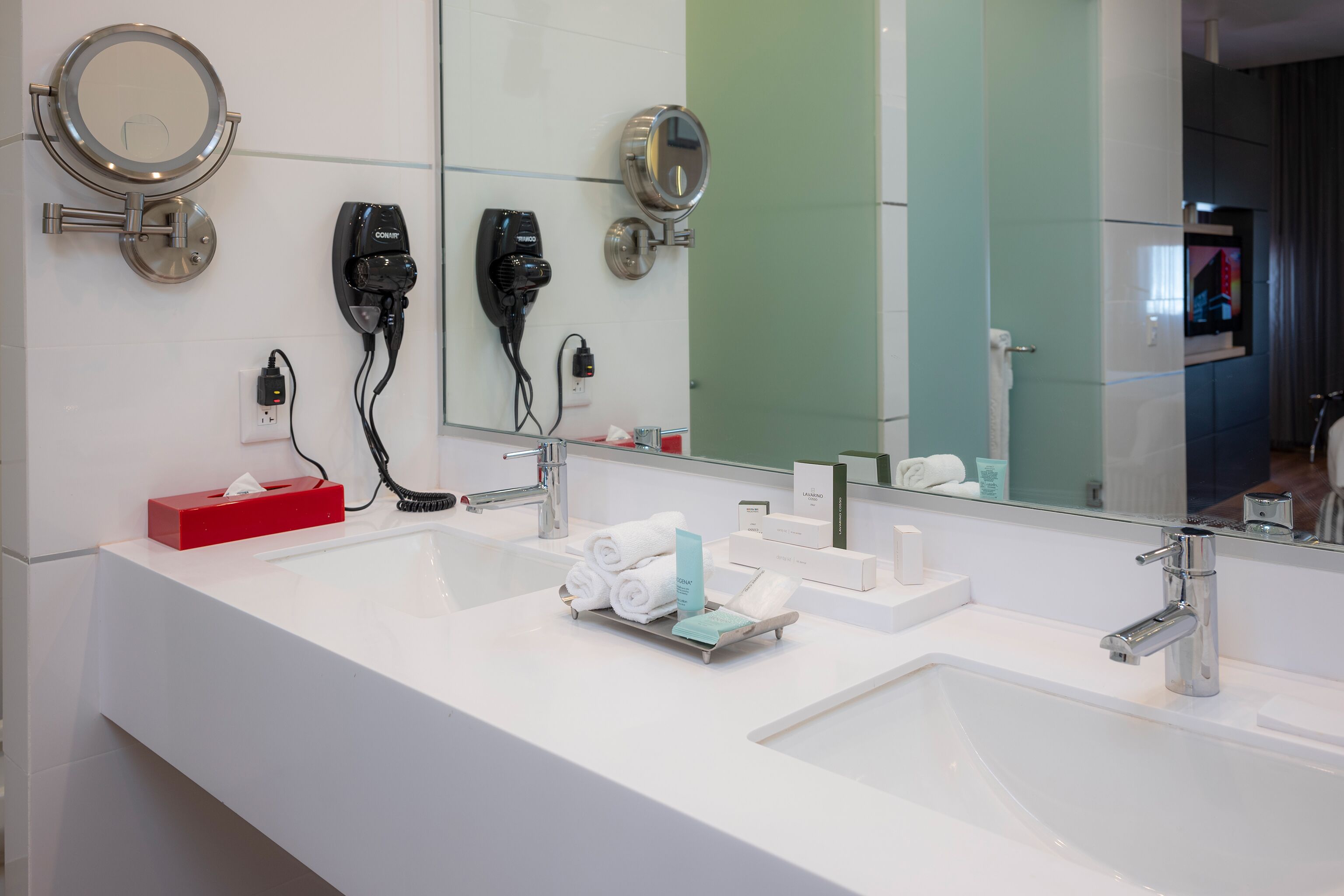 A modern bathroom with two sinks, a large mirror, and hair dryers mounted on the wall.