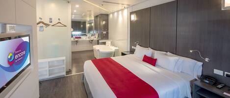 A modern hotel room with a large bed, a bedside table, a TV, and a bathroom visible in the background.