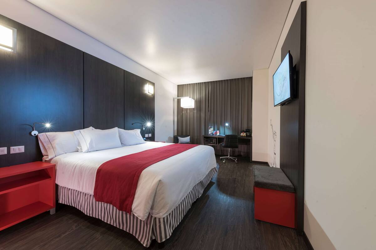 A modern hotel room with a large bed, a desk, a chair, a TV, and a red bench.
