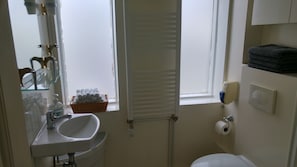 A small bathroom with a white sink, a mirror, a window with blinds, a toilet, and a shelf with towels. - Guesthouse Sólgarðar (Akureyri)