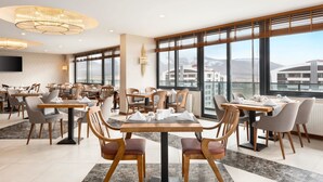 Un espace repas moderne avec des tables et des chaises en bois, de grandes fenêtres offrant une vue sur les montagnes et un téléviseur à écran plat fixé au mur.