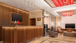 Un espace d’accueil moderne comprenant un comptoir de réception en bois, un téléviseur à écran plat et un coin salon avec des fauteuils et un canapé.