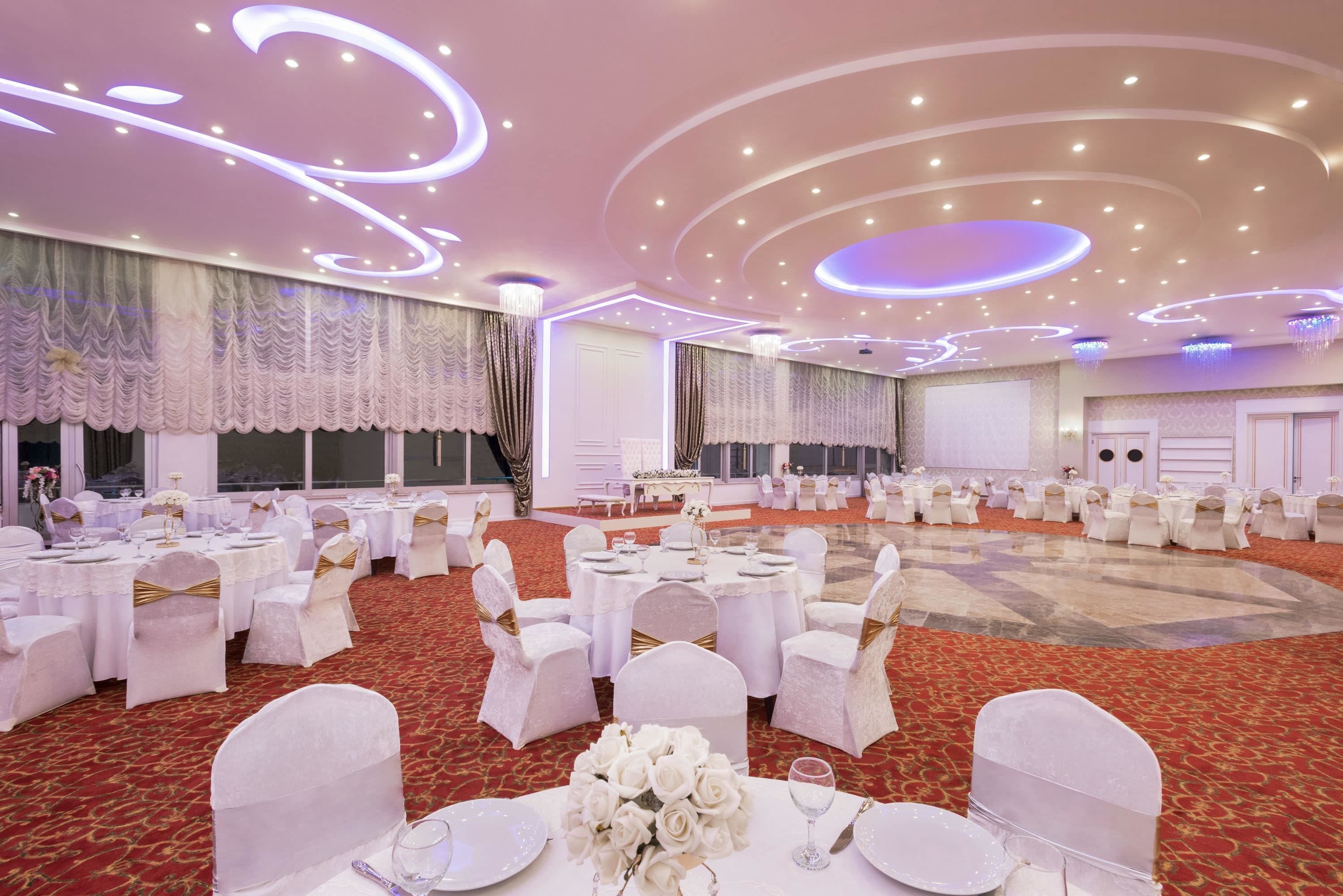A banquet hall with round tables, white chairs, and chandeliers.