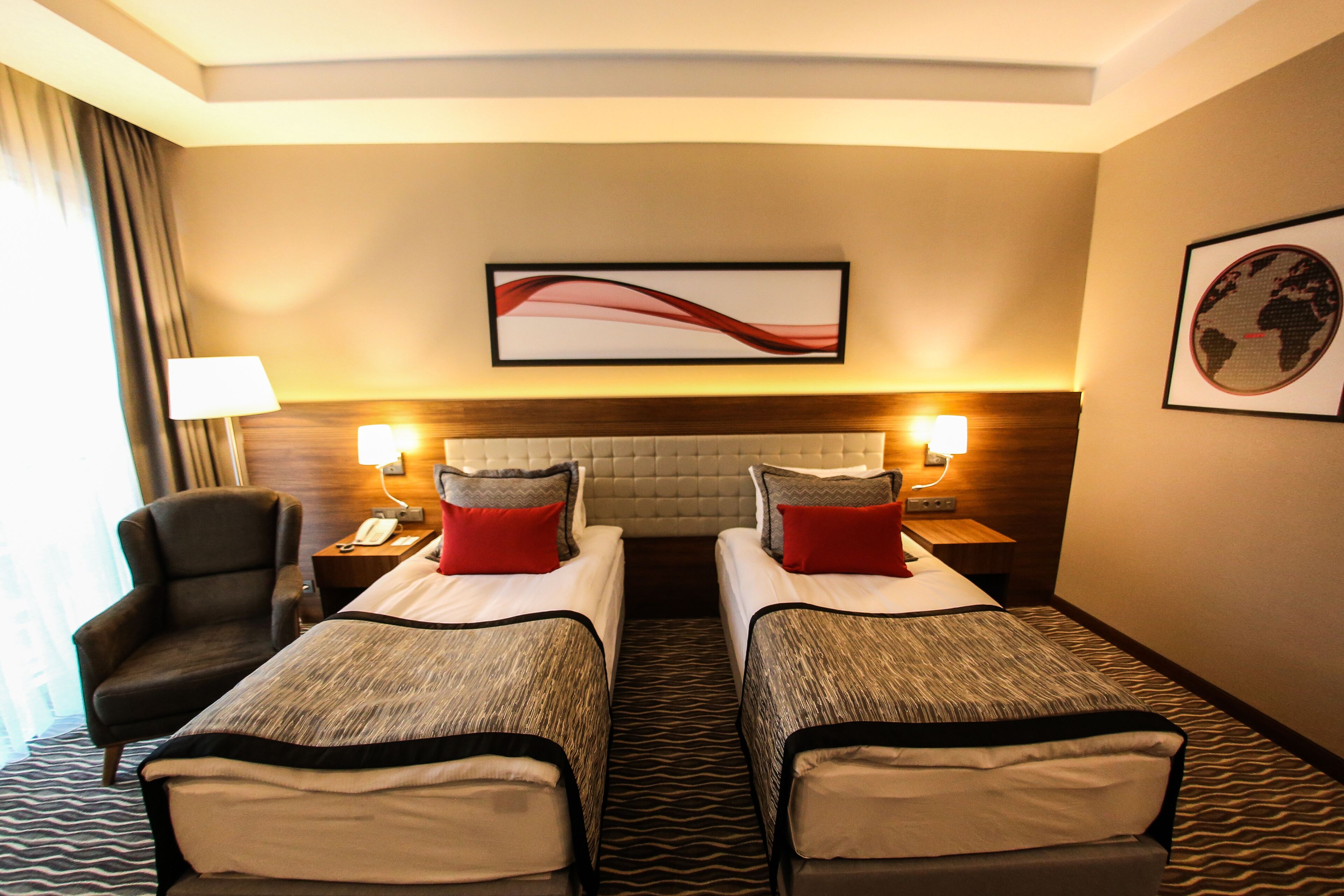 A hotel room with two single beds, a sofa, a chair, a desk with a lamp, and a world map on the wall.