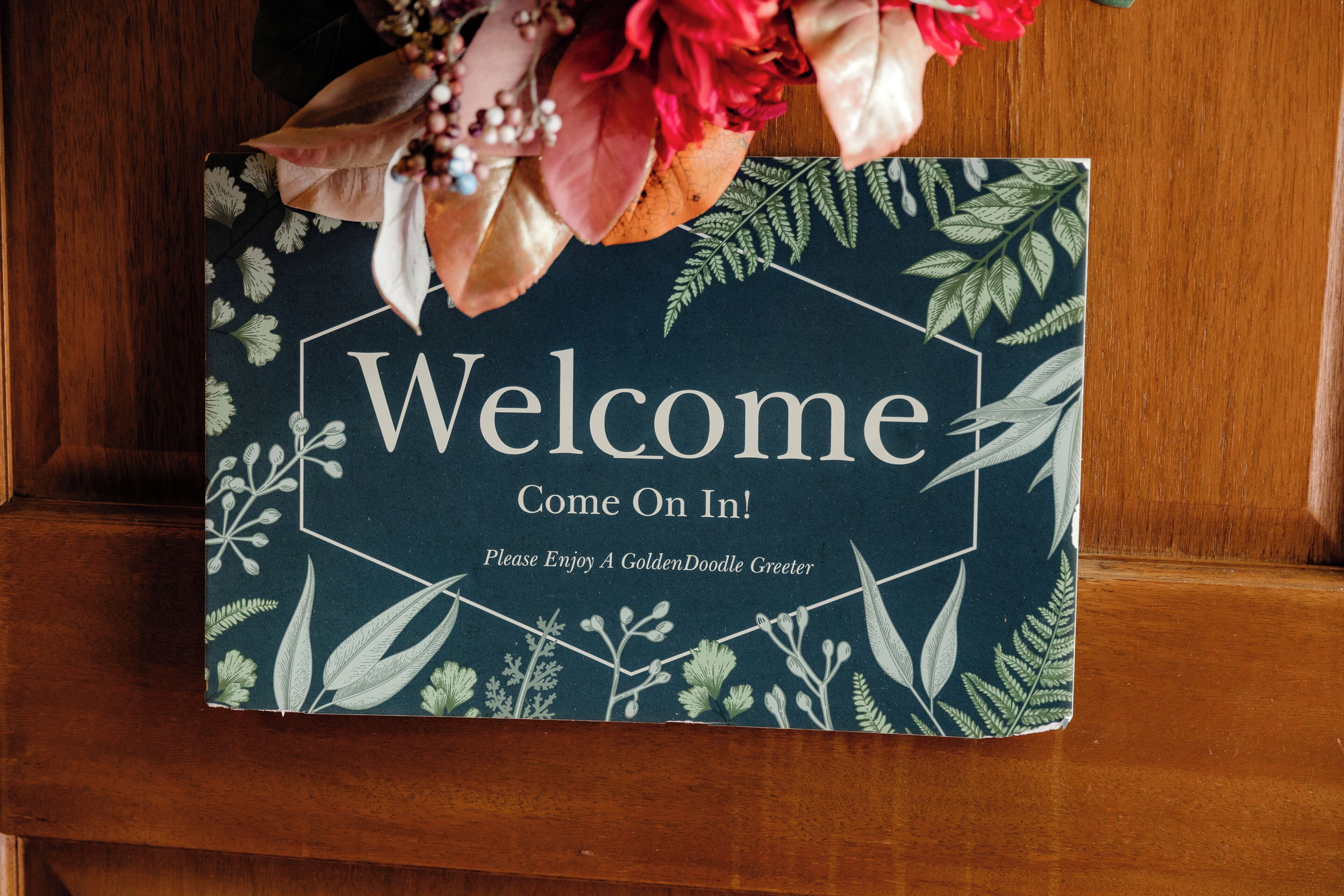 A welcome sign with a floral pattern and the text 'Welcome, Come On In! Please Enjoy A GoldenDoodle Greeter.'