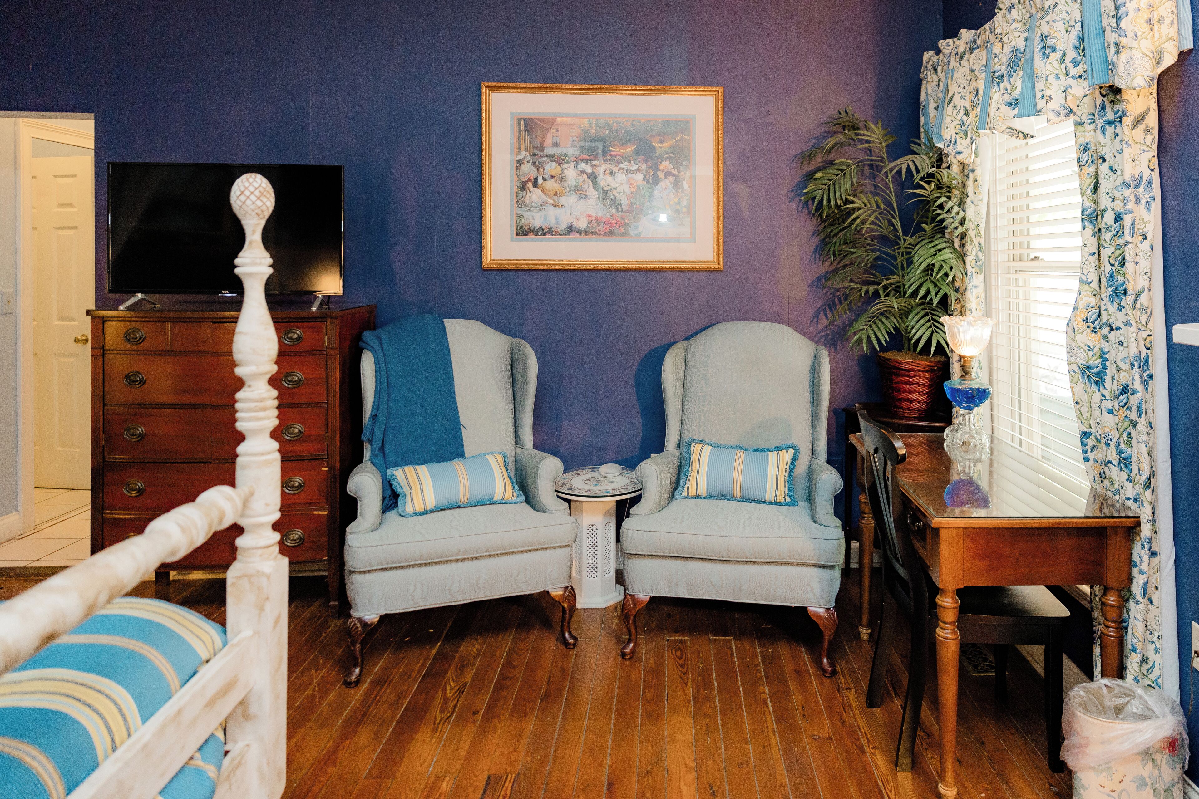 A room with a wooden floor, two armchairs with blue cushions, a small round table, a dresser, a television, a framed picture on the wall, a potted plant, and a window with curtains.