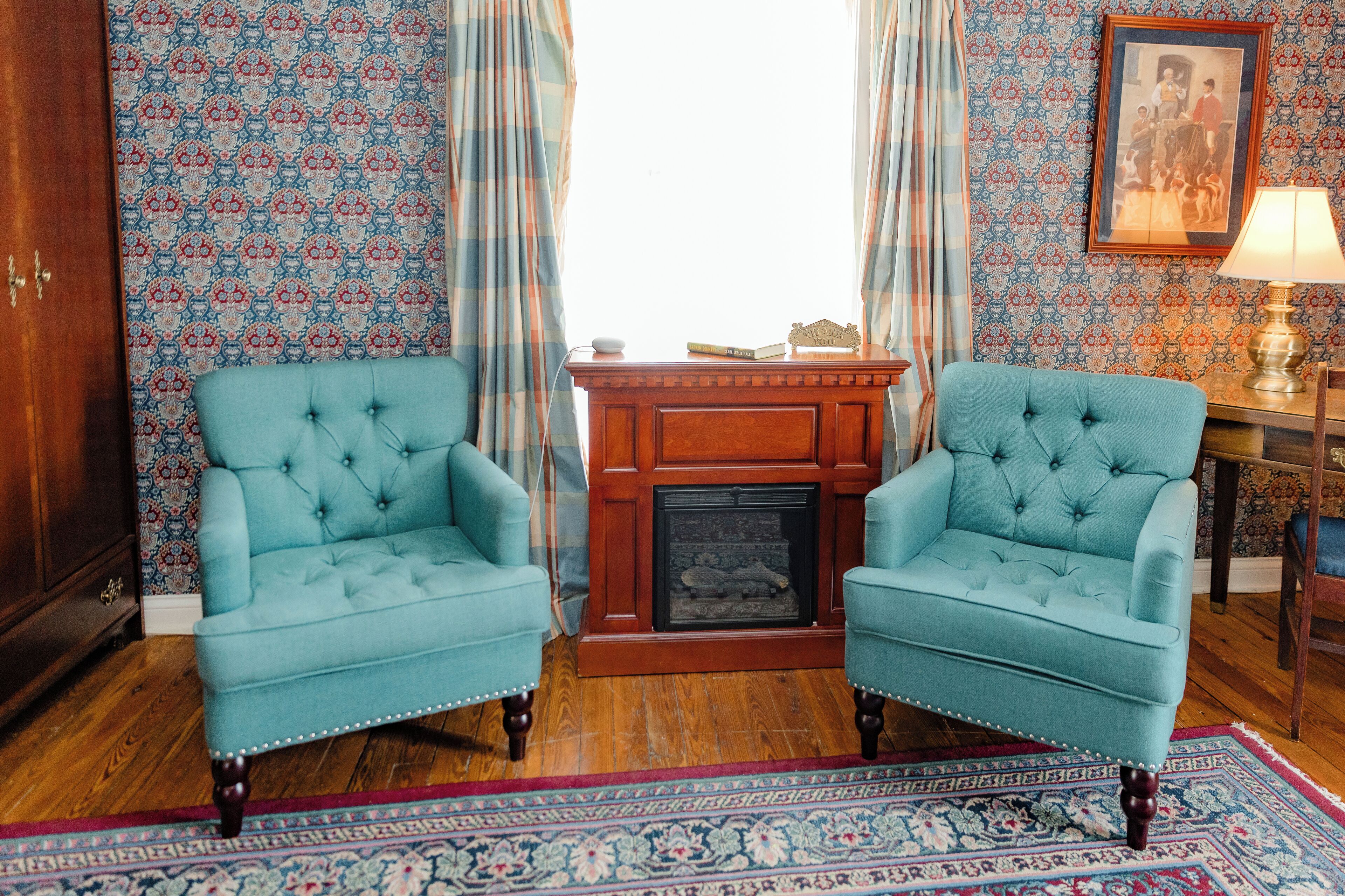 A room with two upholstered armchairs, a wooden sideboard, a fireplace, and patterned wallpaper.
