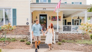 A couple holding hands in front of a house with a porch, a stone wall, and an American flag. - Montgomery Inn BnB (Versailles)