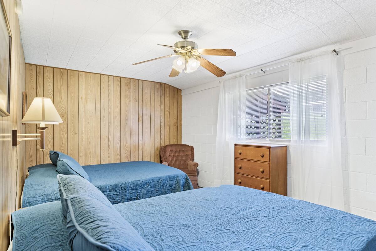 Ein Schlafzimmer mit zwei Betten, einem Deckenventilator, einer Holzwand, einem Fenster mit Jalousien und einem Schminktisch.