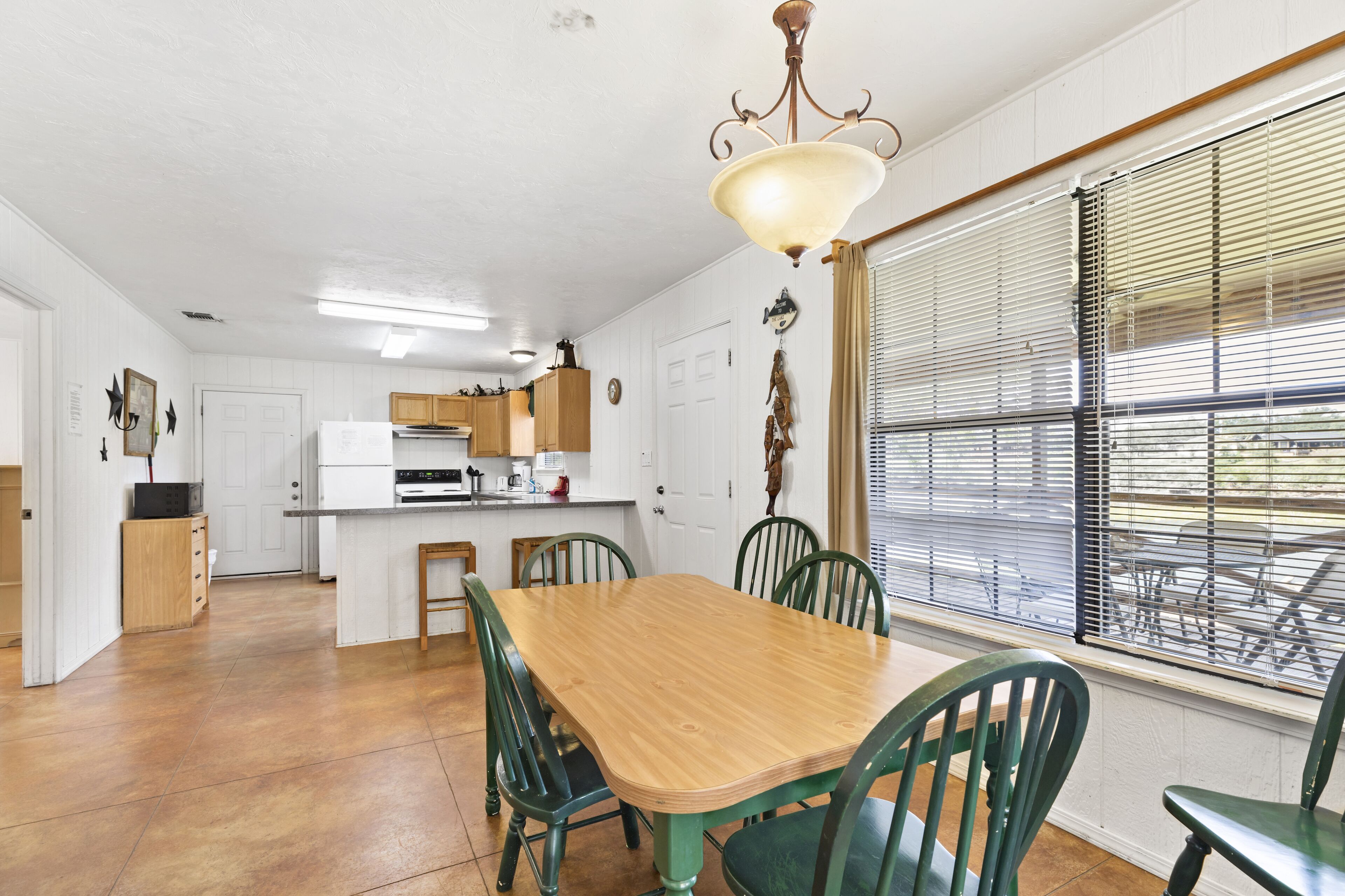 Un comedor con una mesa de madera y sillas verdes, una cocina al fondo y una ventana con persianas.