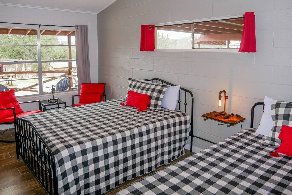 Ein Zimmer mit zwei Betten, roten Kissen, einem kleinen Tisch mit Lampe und einem Fenster mit Vorhängen.