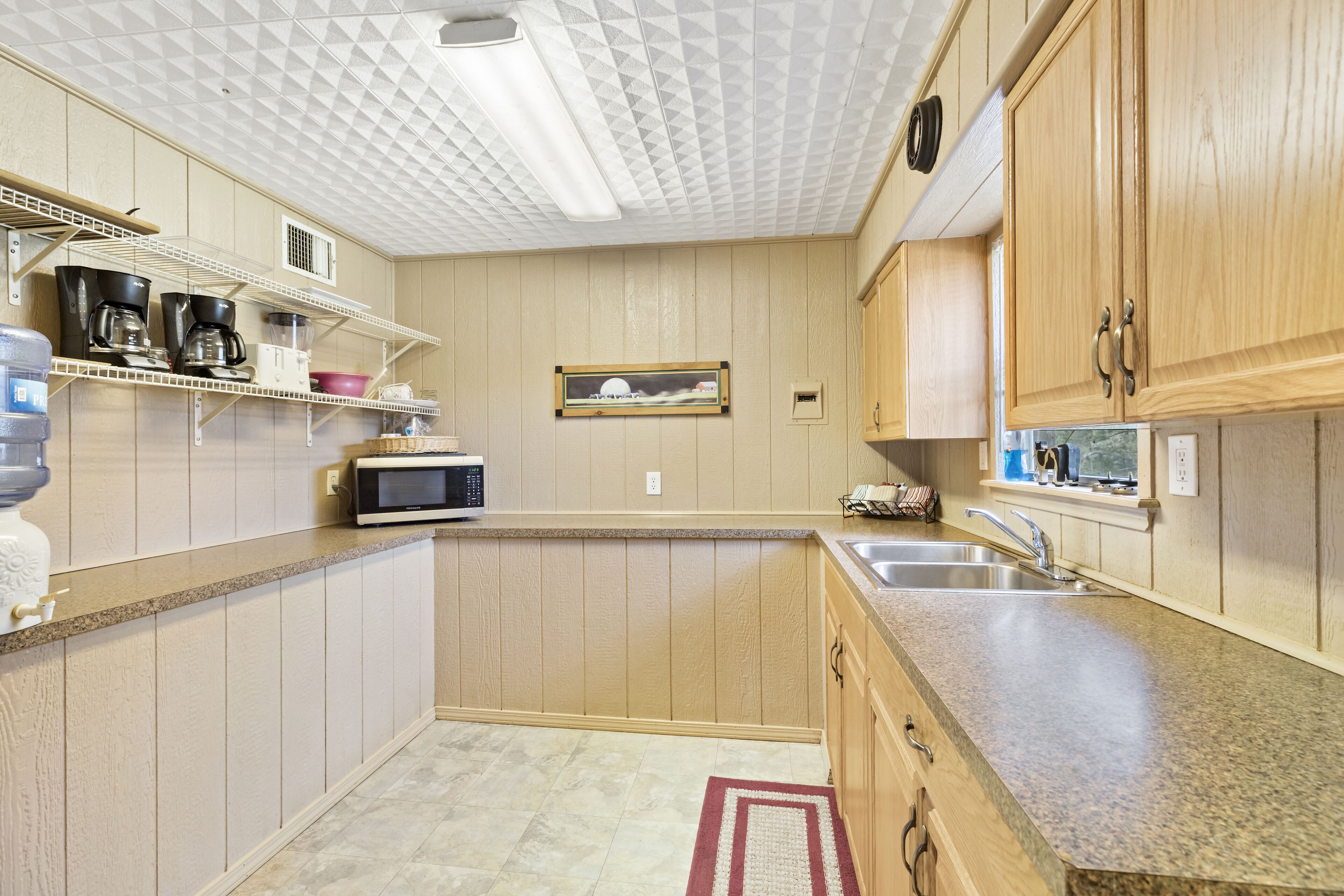 A kitchen with wooden cabinets, a microwave, a sink, and a shelf with kitchen items.