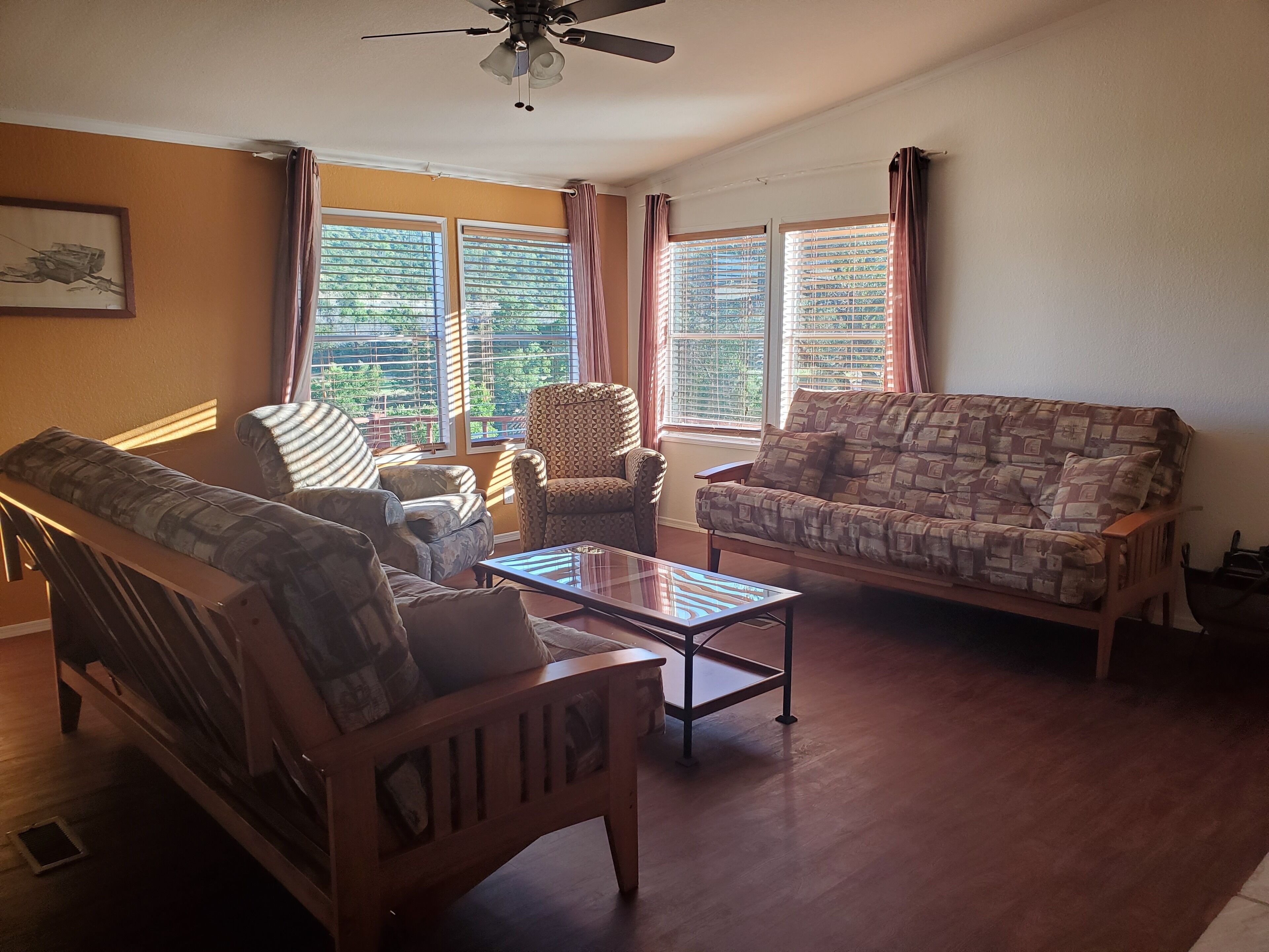 A living room with a sofa, armchair, and a coffee table. There are windows with curtains, a ceiling fan, and a framed picture on the wall.