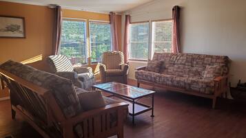 A living room with a sofa, armchair, and a coffee table. There are windows with curtains, a ceiling fan, and a framed picture on the wall.