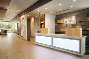 A hotel reception area with a modern design, including a reception desk, a wall-mounted sign for Holiday Inn Express & Suites, and a large window providing natural light.
