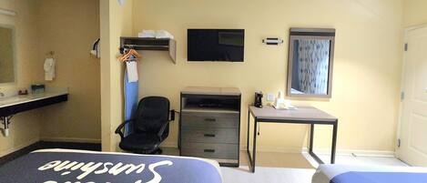 A hotel room with two beds, a desk, a chair, a TV, a mirror, and a small shelf with items.