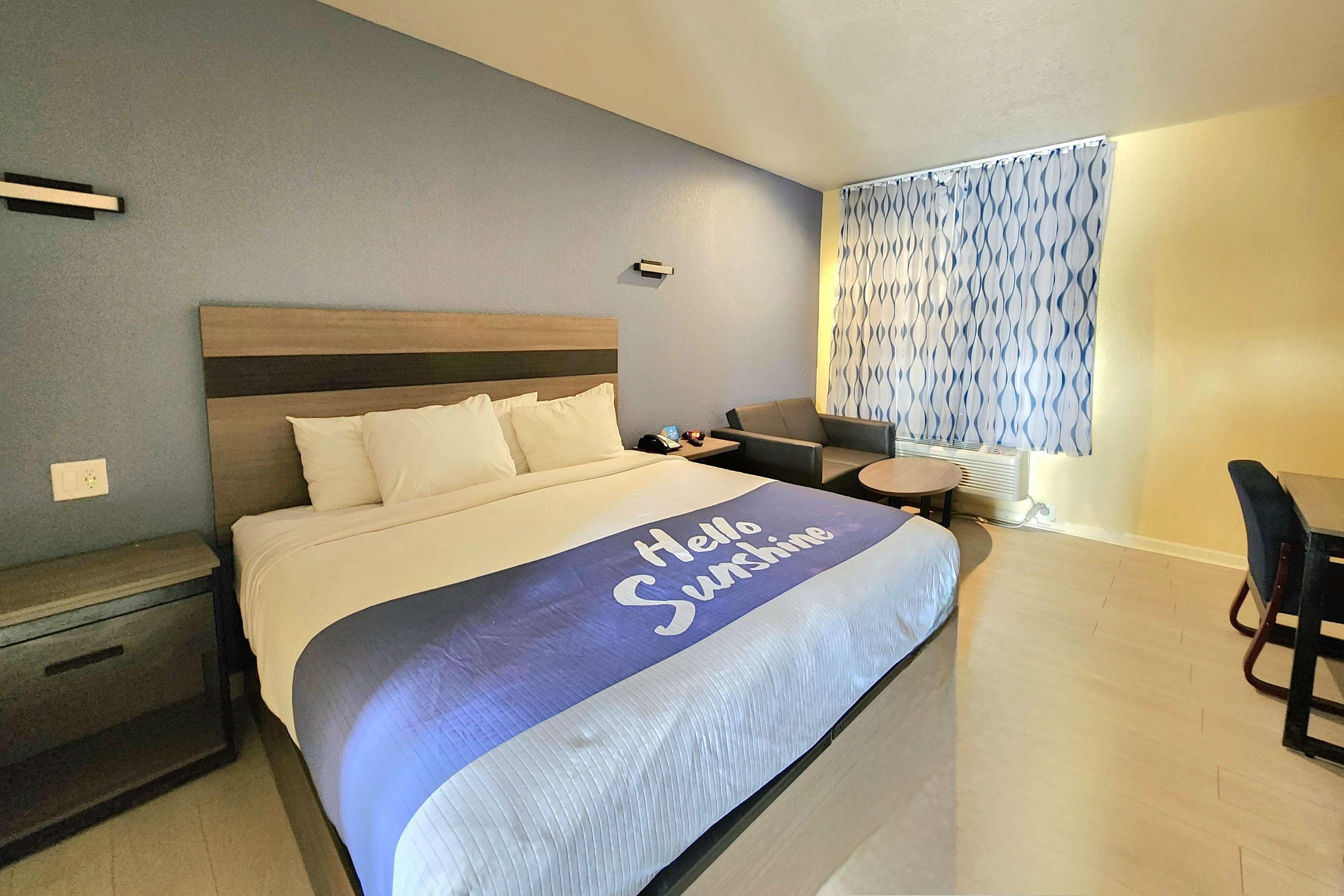 A neatly made bed with a "Hello Sunshine" pillow, a bedside table, a chair, and a small round table.