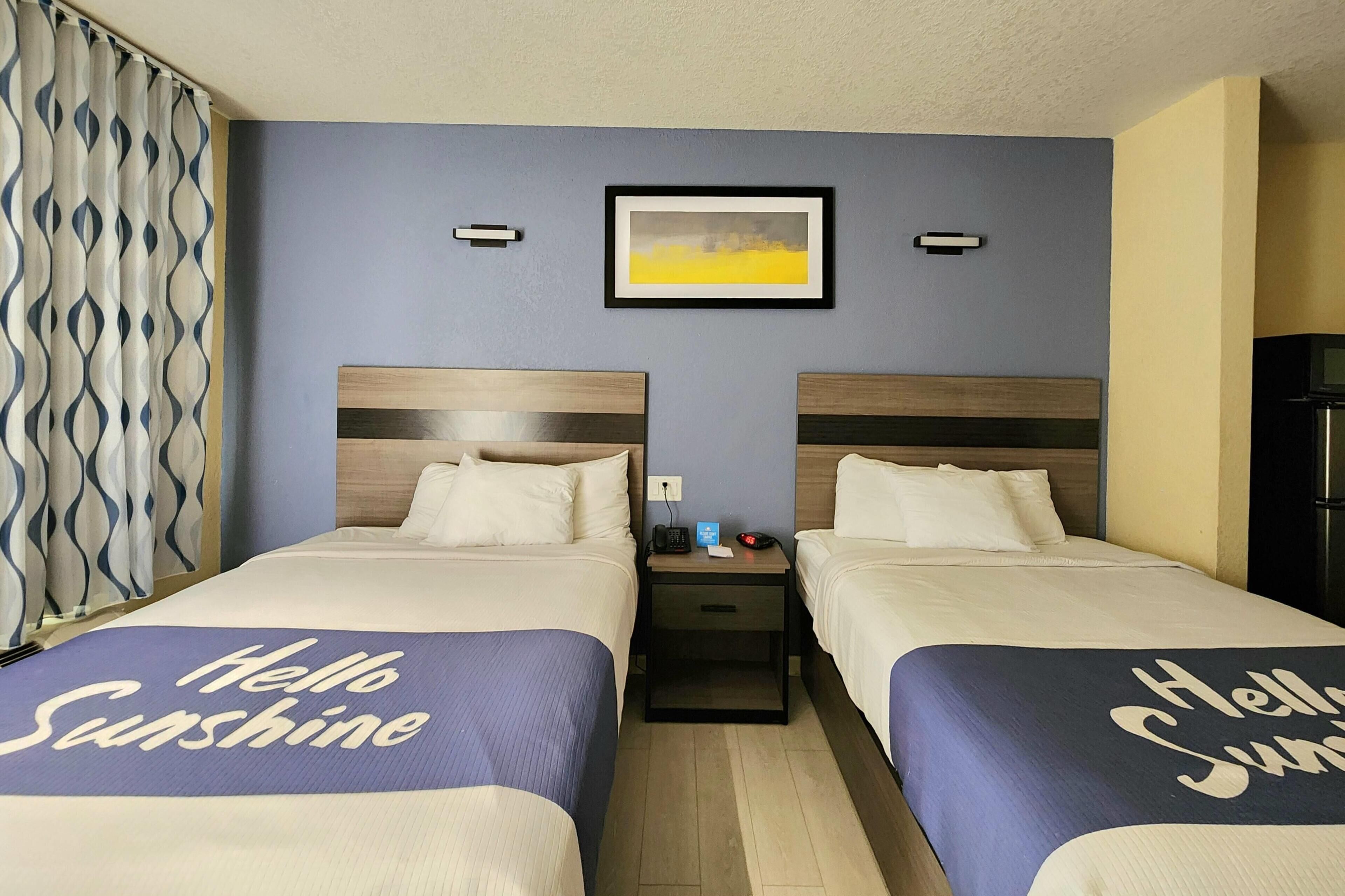 Una habitación de hotel con dos camas, una mesita de noche con un teléfono y una lámpara, un cuadro en la pared y una cortina con un estampado ondulado.