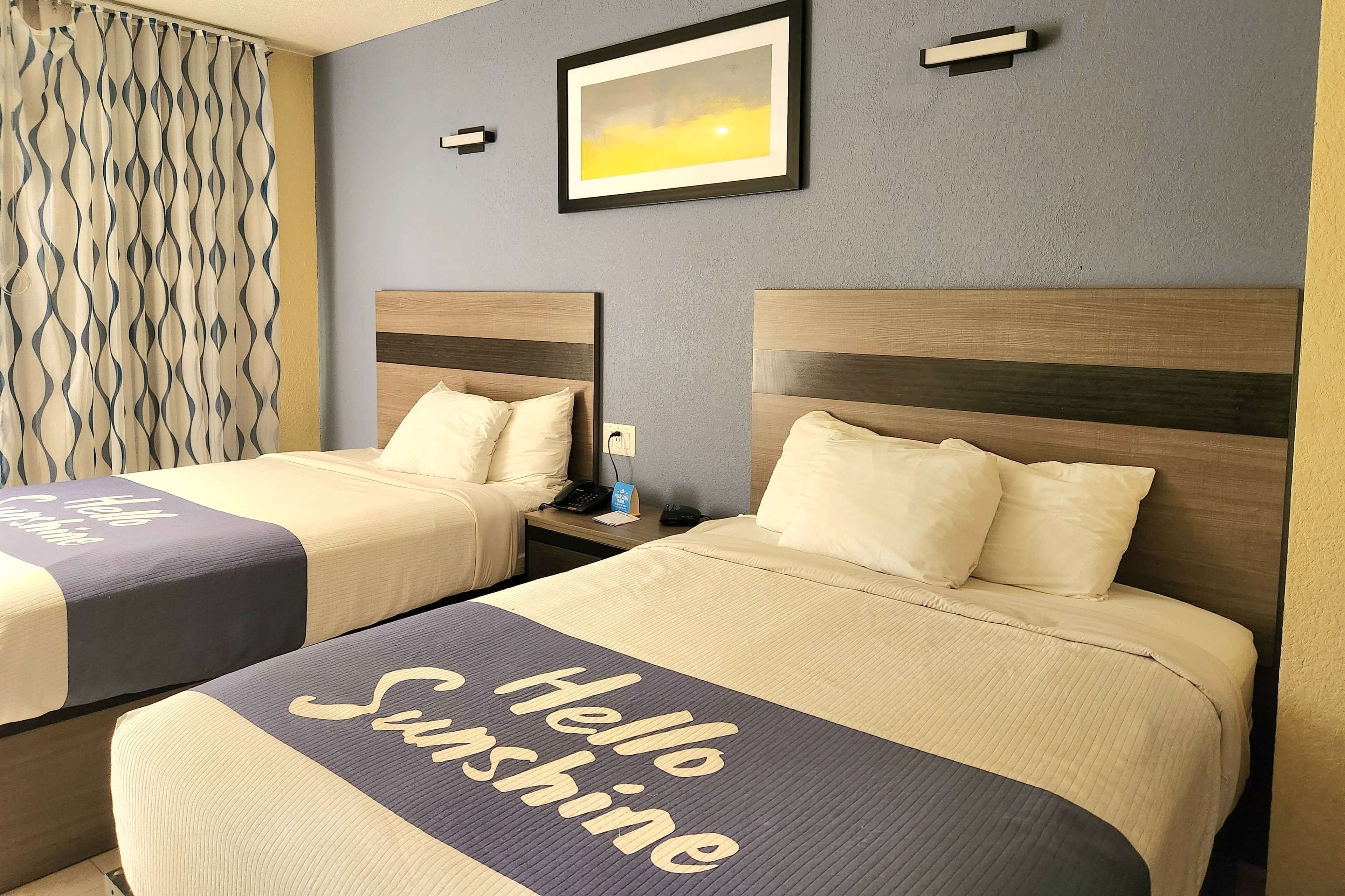 A hotel room with two beds, each with a headboard and a pillow. There is a framed picture on the wall, a curtain with a wavy pattern, and a bedside table with a phone and a small lamp.