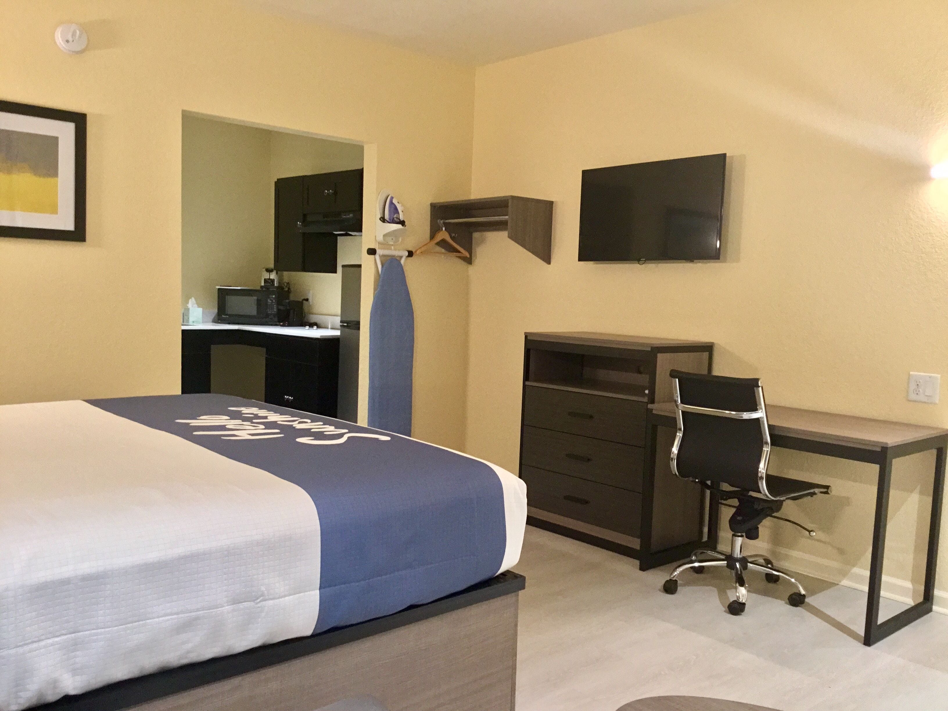 Un dormitorio con cama, escritorio, silla, televisor y una kitchenette al fondo.