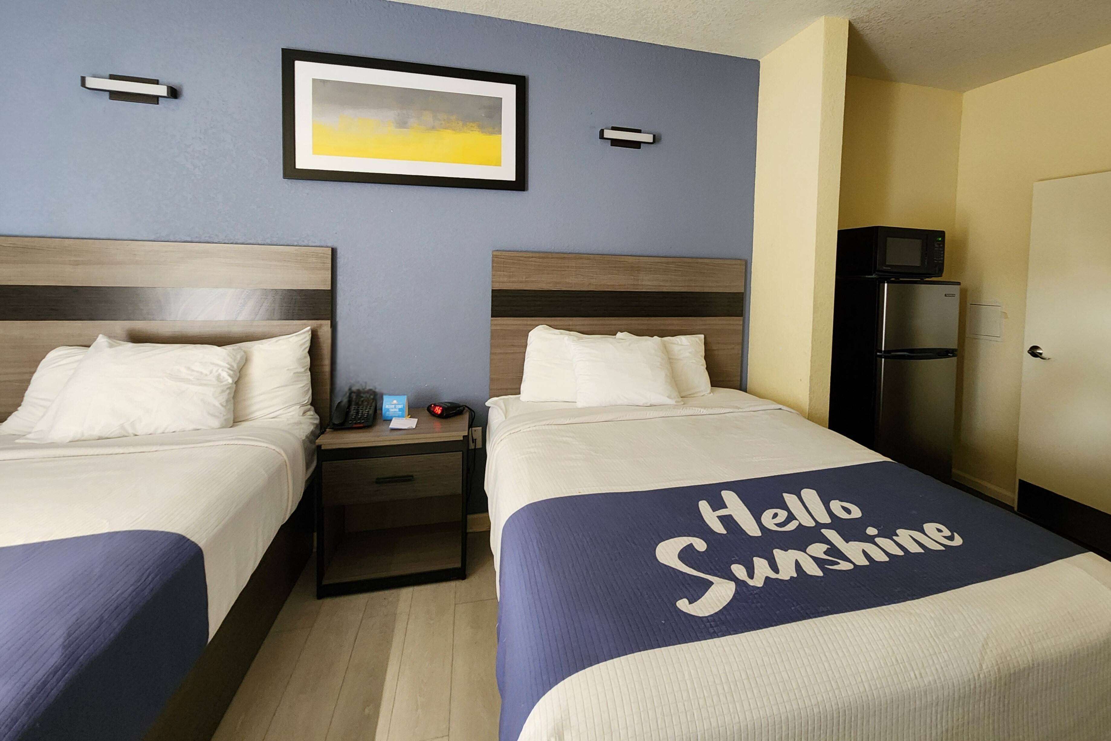 Una habitación de hotel con dos camas, una mesita de noche, un cuadro enmarcado en la pared, un microondas y un refrigerador.