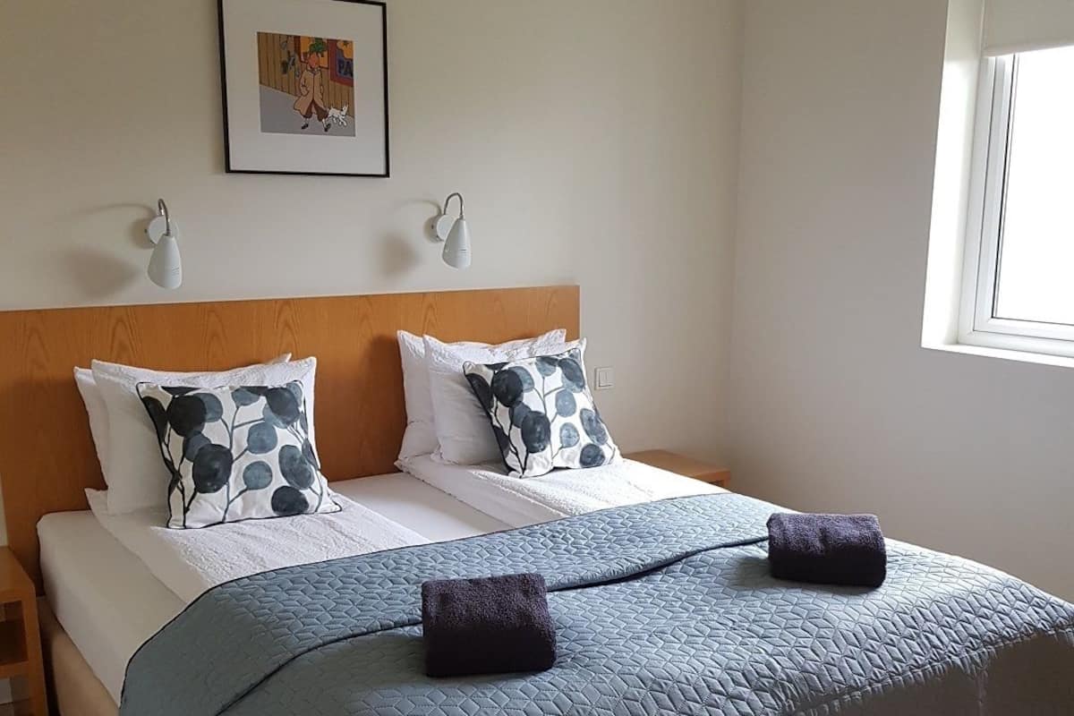 A neatly made bed with a blue quilt and two decorative pillows, a wooden headboard, a framed picture on the wall, and a window with blinds.