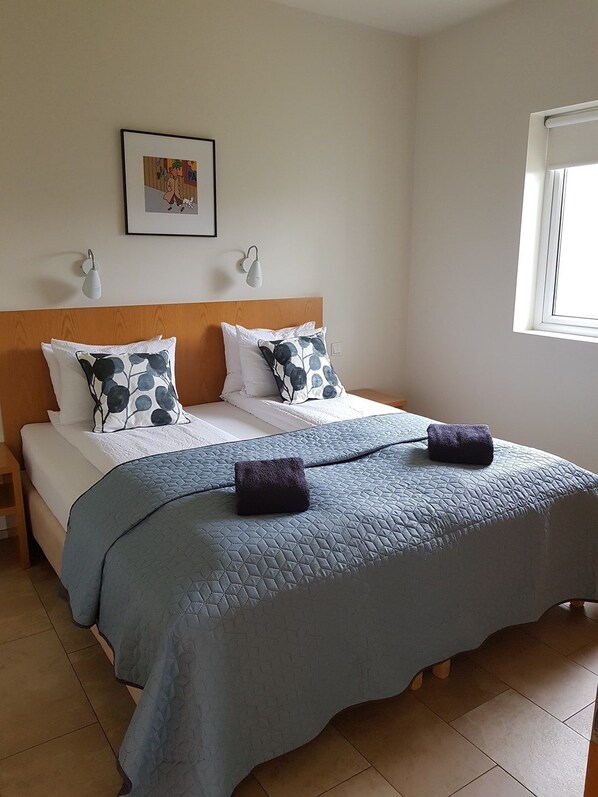 A neatly made bed with a blue quilt and two decorative pillows, a wooden headboard, a framed picture on the wall, and a window with blinds. - Sæluhus Hotel Apartments & Houses (Akureyri)