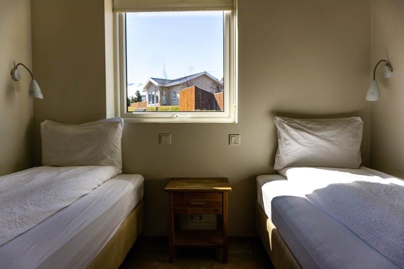 Een kleine, eenvoudige kamer met twee bedden, een raam en een nachtkastje.