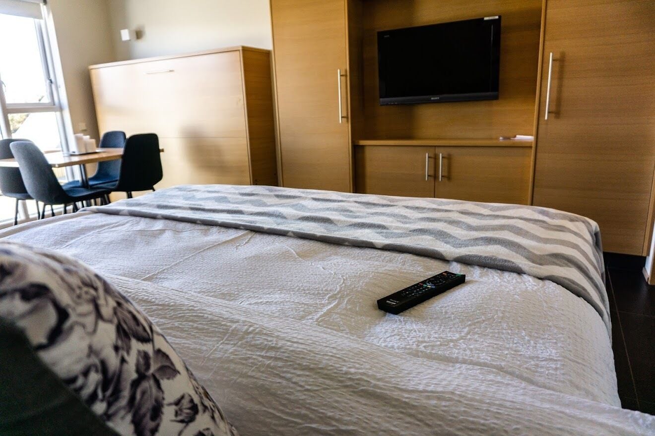 A hotel room with a bed, a TV mounted on the wall, a desk with chairs, and wooden cabinets.