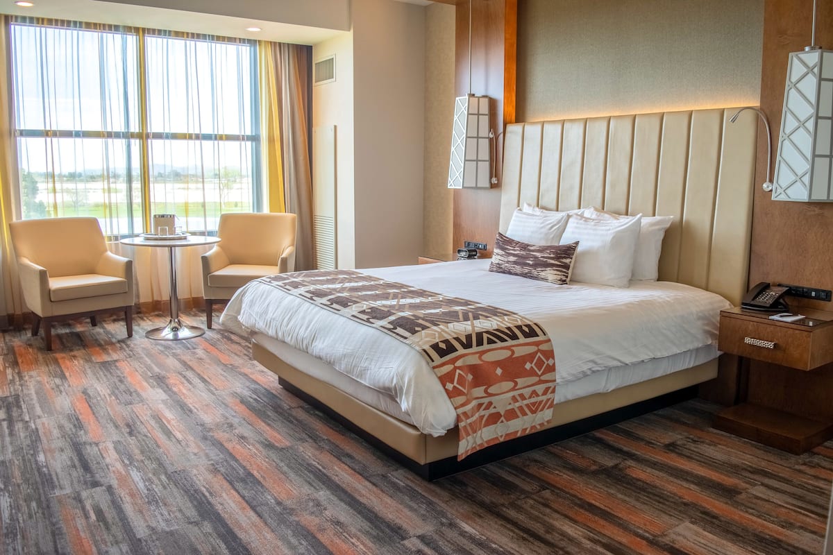 Habitación de hotel con una cama grande, dos sillones, una mesita y vistas al exterior a través de amplias ventanas.