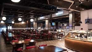 Un restaurant spacieux doté de nombreuses tables et banquettes, d’un comptoir où un chef prépare les plats, et d’un décor intérieur moderne agrémenté de motifs géométriques sur les murs et le sol.
