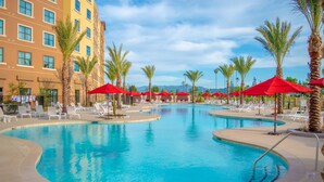 Un espace piscine d’hôtel avec de nombreux chaises longues et des parasols rouges, entouré de palmiers et d’un immeuble à plusieurs étages.