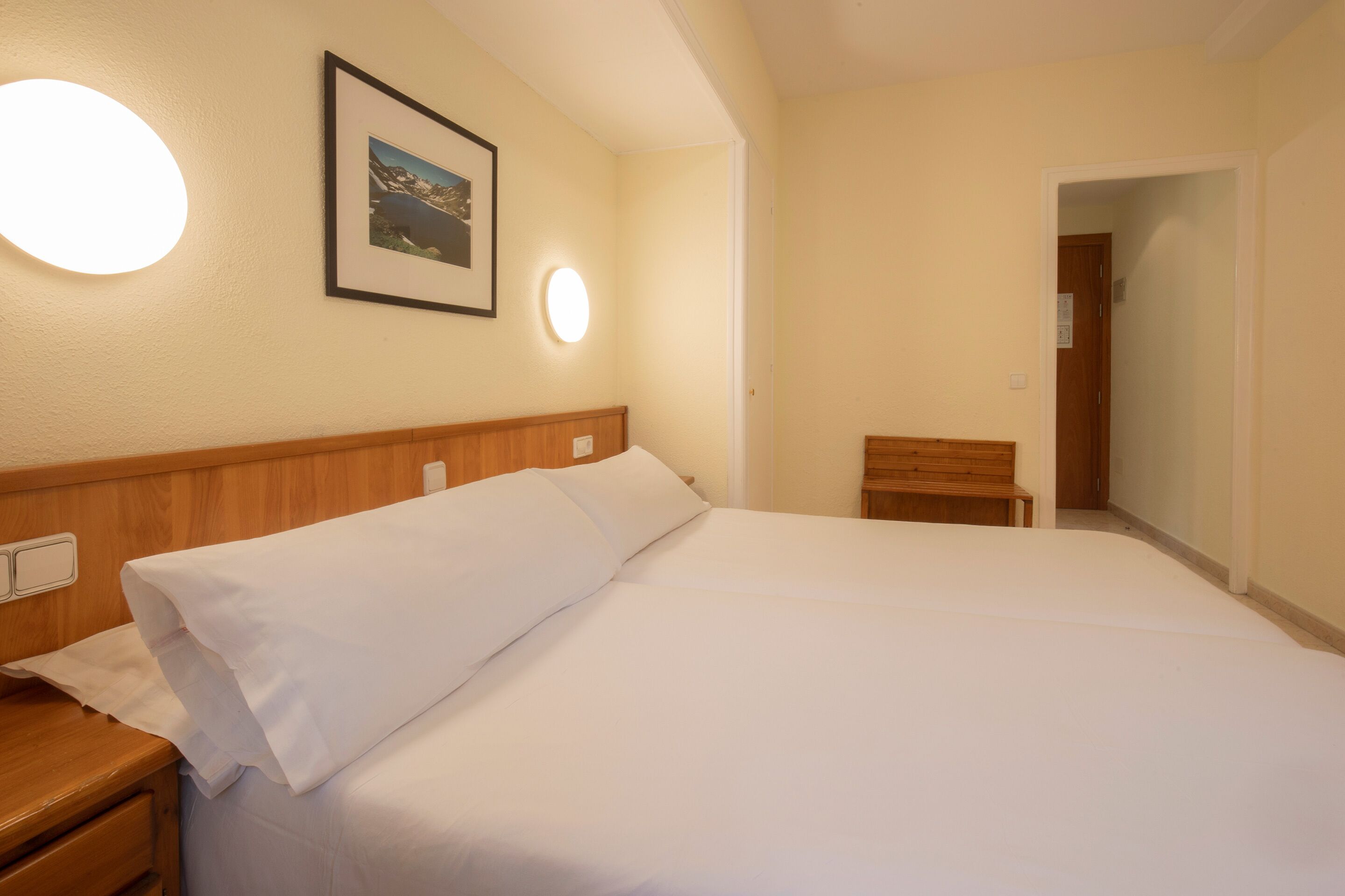 A neatly made bed in a hotel room with a wooden headboard, a framed picture on the wall, and a small wooden bench.