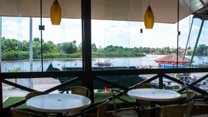 Un espace extérieur aménagé avec des tables et des chaises en osier, offrant une vue sur une rivière où naviguent des bateaux et sur un bâtiment situé au loin.