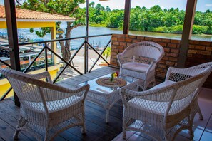 Un balcón con mobiliario de mimbre y una mesa de vidrio, con vistas a un río y vegetación exuberante.