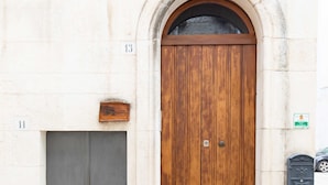 A building with a large wooden door and a smaller grey door, both with metal handles. There is a small window above the larger door and a mailbox on the right side of the building. - B&B La Stradella (Ostuni)