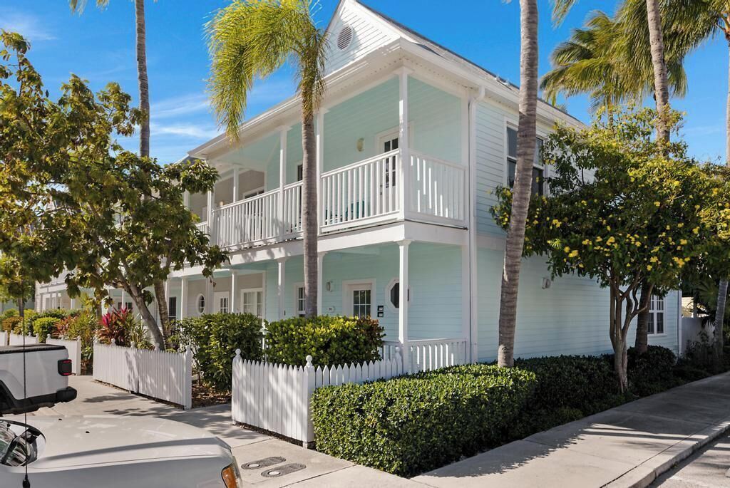 A two-story house with a balcony, surrounded by palm trees and a white picket fence.