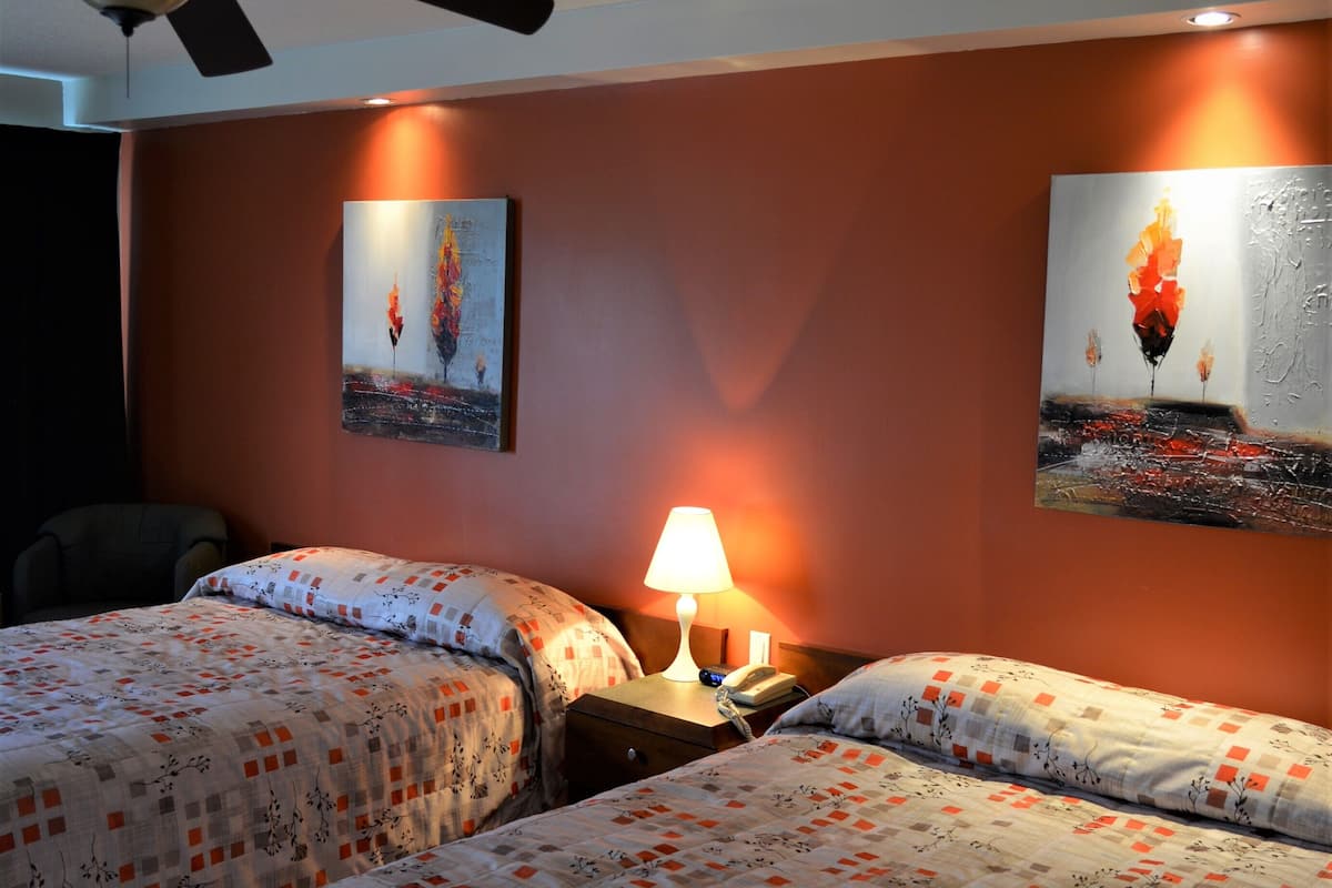 A hotel room with two beds, a ceiling fan, a nightstand with a lamp, and two paintings on the wall.