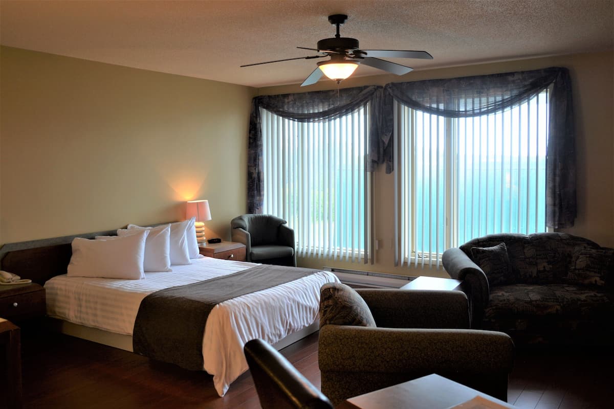 A hotel room with a bed, two armchairs, a table, a desk, a lamp, a phone, and a ceiling fan.
