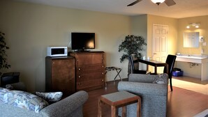 A living room with a flat-screen TV on a wooden cabinet, a ceiling fan, a dining table with chairs, a potted plant, and a bathroom with a sink and mirror. - Motel Quatre Saisons (Forestville)
